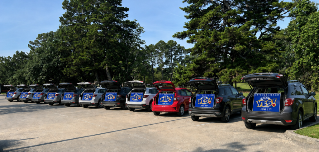 Line of vehicles parked with their rear hatches open, displaying "Vote YES! Save Our Library" signs during an East Baton Rouge Parish Library advocacy campaign.